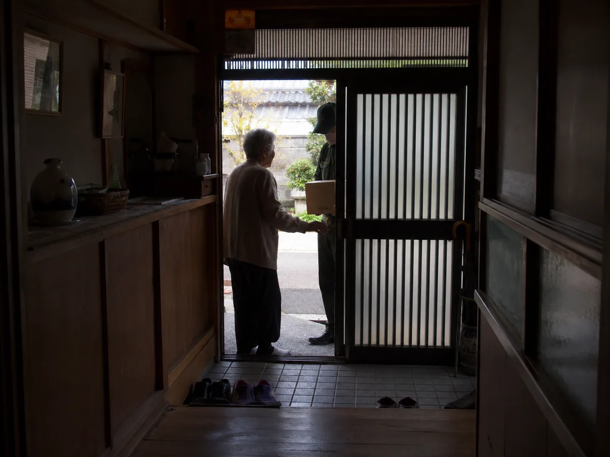 玄関先で配達員から荷物を受け取り言葉を交わす一人暮らしの高齢女性