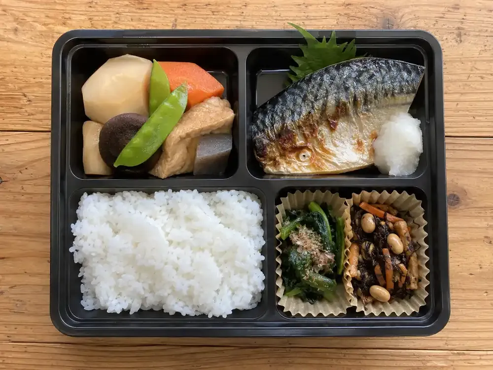 焼き魚と煮物、青菜のおひたし、ご飯が入った配食サービスの弁当