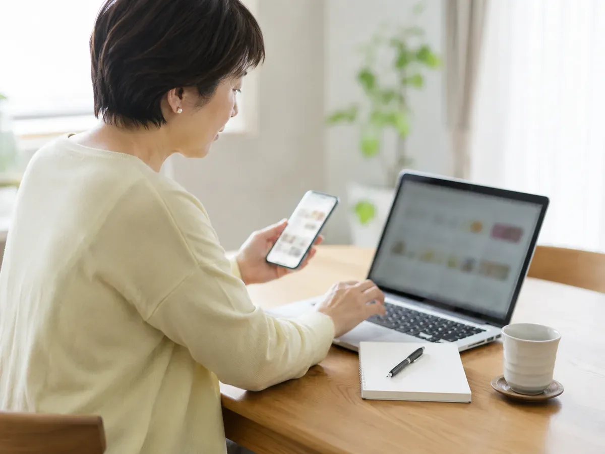 ノートパソコンとスマートフォンで複数のネットスーパーを見比べる女性